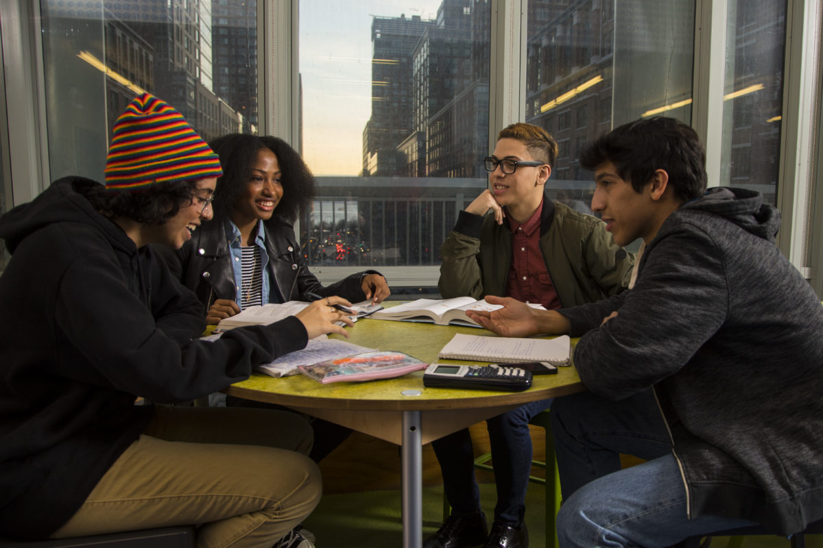 Four students gathered around a table with books and calculators, talking and laughing