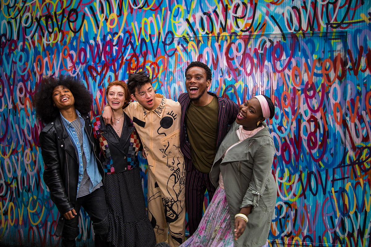 A group of smiling young people, laughing and smiling in front of a wall that says “love”