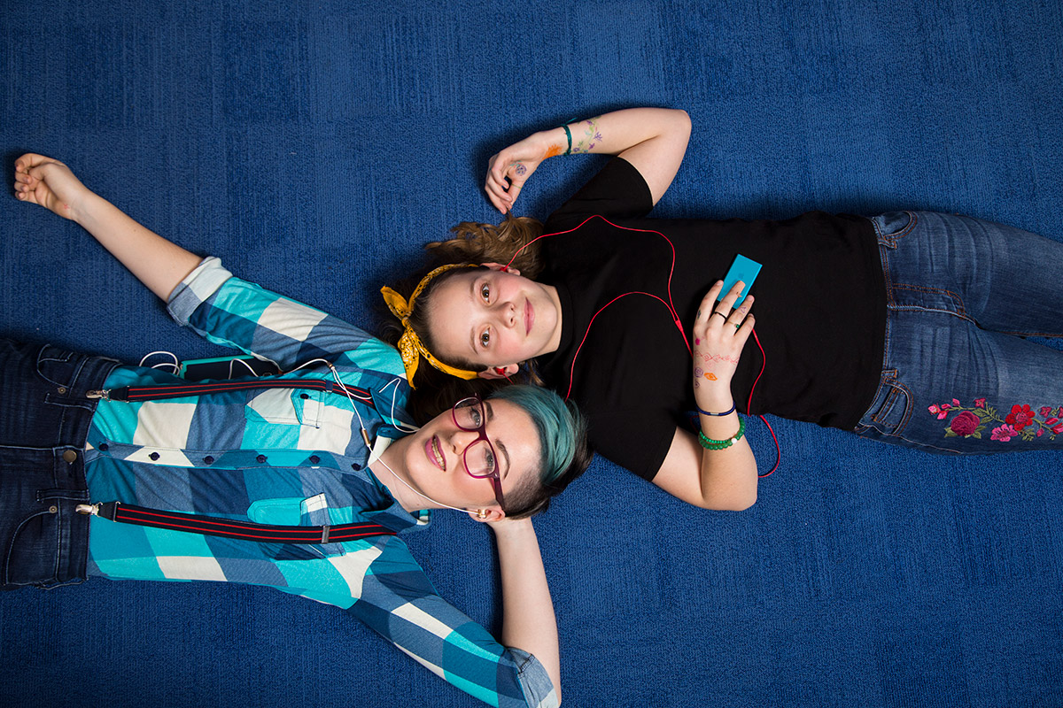 Two young people lying head-to-head on blue carpet, both with headphones in their ears