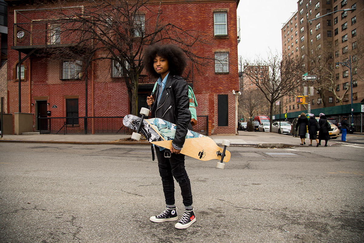 A young person standing on the street in front of a brick building carrying a skate board and a backpack