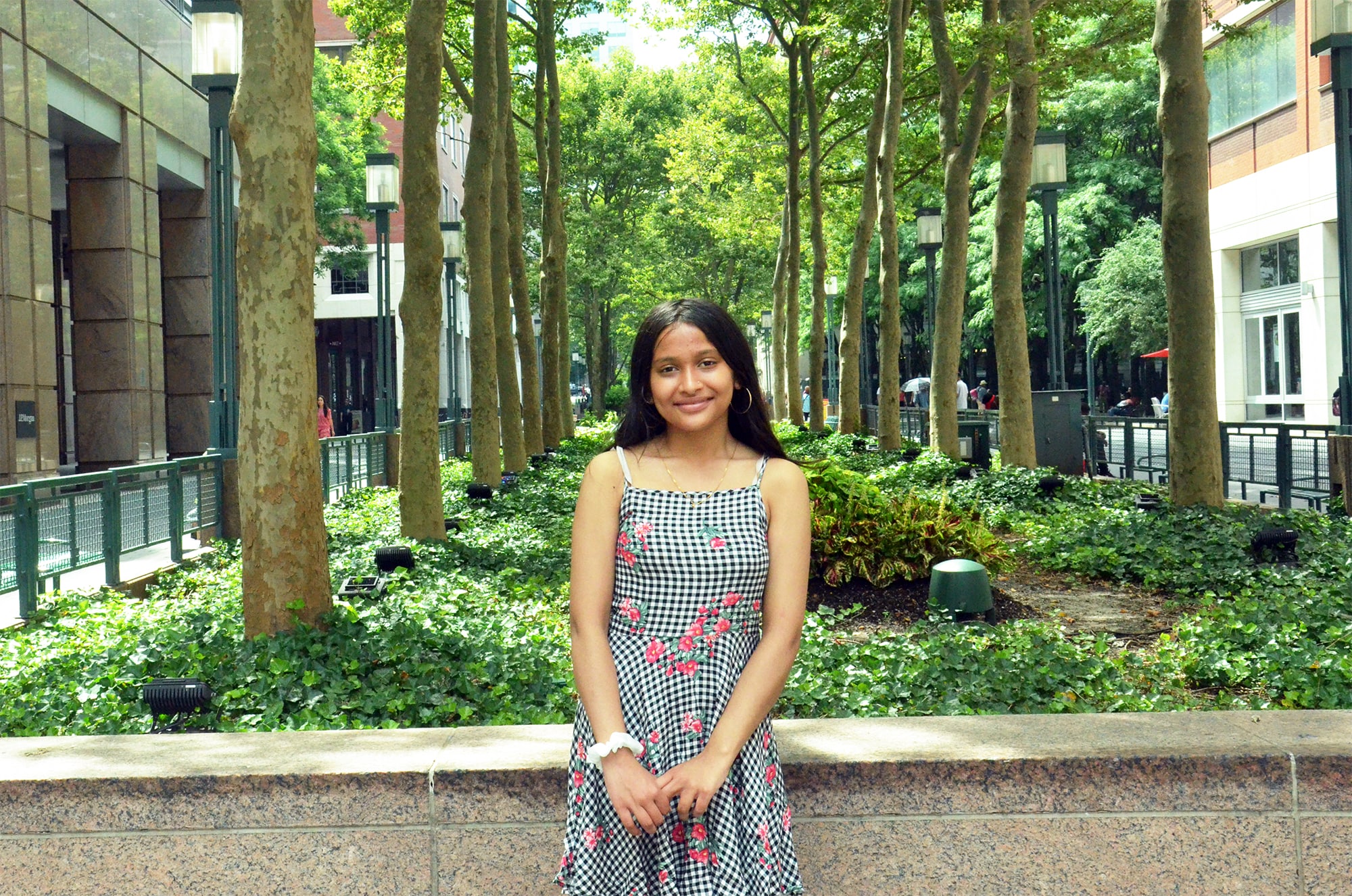 A teenager wearing a checked dress poses in front of a row of trees at Metrotech Center in Brooklyn