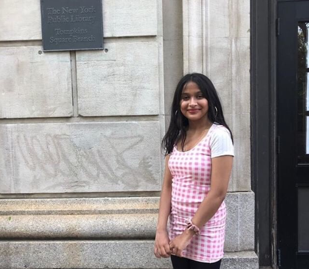 A teenager wearing a pink dress smiles in front of the Tompkins Square Branch of the New York Public Library