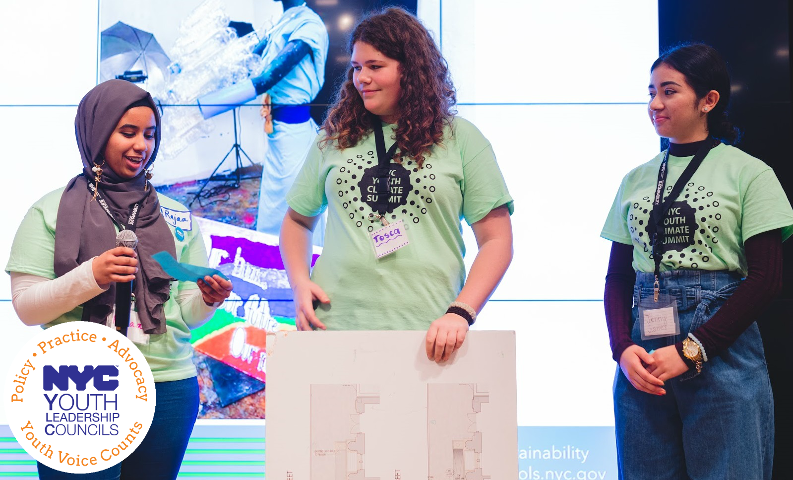 Three teenagers in matching green tee-shirts stand on stage presenting a poster board